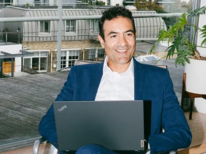 A man sitting outdoors with a laptop, smiling at the camera.